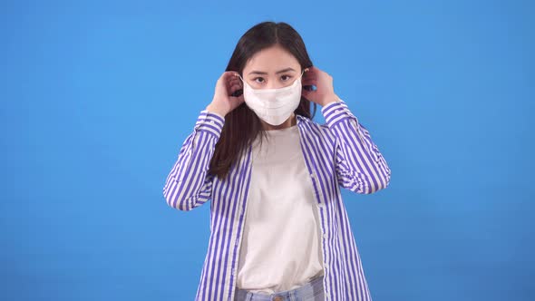 Beautiful Young Asian Woman Puts a Protective Medical Mask on Her Face Standing on a Blue Background alt