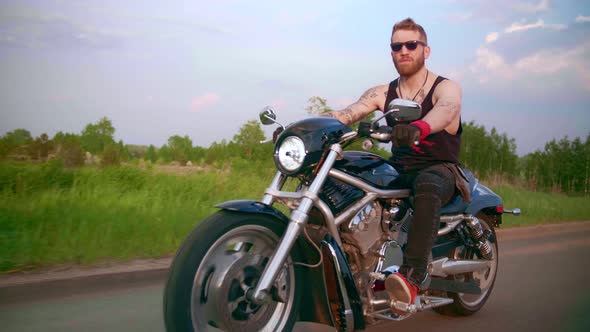 Stylish Biker with Tattoos Rides a Motorcycle on a Country Road at Sunset