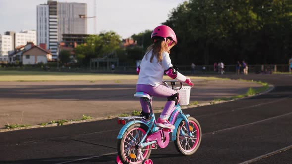 Cute Light Hair Little Girl in Pink Helmet in Elbow and Knee Pads Rides a Bicycle at the Stadium