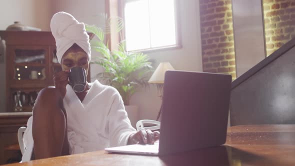 African american attractive woman with applied face mask drinking coffee and using laptop at home alt