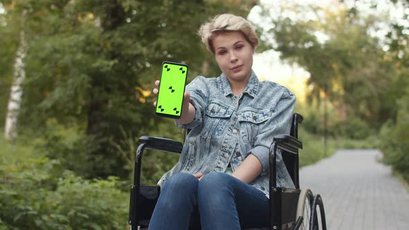 Disabled Woman is Sitting in Wheelchair in Park and Shows Phone with Chromakey Mockup alt
