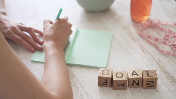 Girl At The Table And Writes In Notebook. Young Woman Sets Goal To Lose Weight. Word Goal On Table alt