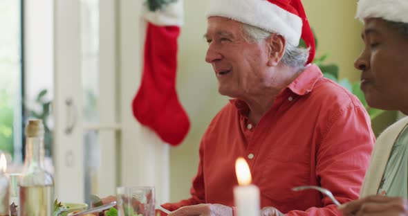 Diverse senior man and woman in santa hats listening and laughing at christmas dinner table at home alt