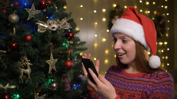 Young Cheerful Happy Woman in Christmas Santa Hat Using Mobile Phone alt