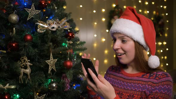 Young Cheerful Happy Woman in Christmas Santa Hat Using Mobile Phone