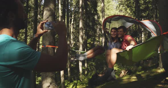 Man Making Smartphone Photo to Smiling Woman on Hanging Tent Camping in Sunny Forest
