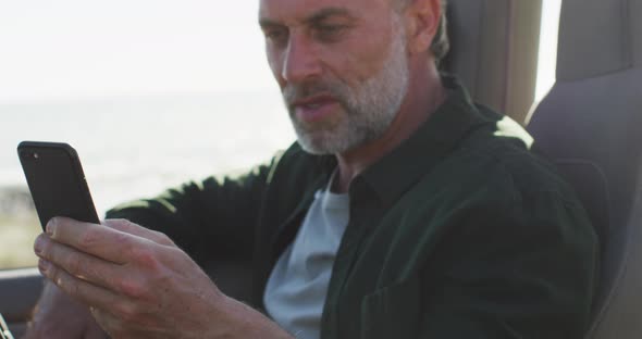Happy caucasian man sitting in car using smartphone on sunny day by the sea alt