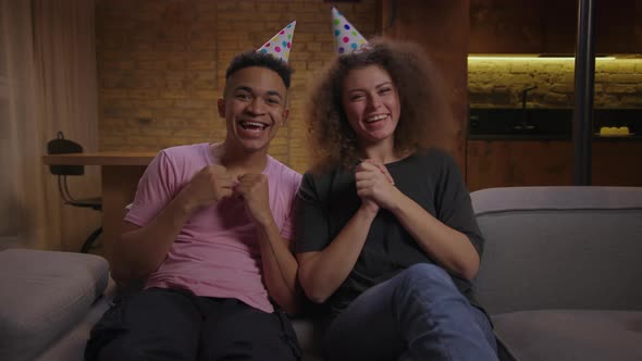 Young Mixed Race Couple in Birthday Hats Wishing All the Best Talking at Camera Sitting on Couch at