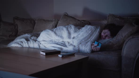 Happy Woman Lies on the Sofa at Night Near the TV and Uses the Phone