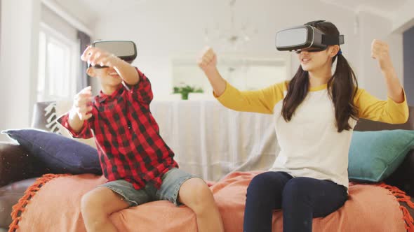Happy asian brother and sister at home, sitting on couch in living room wearing vr headsets alt