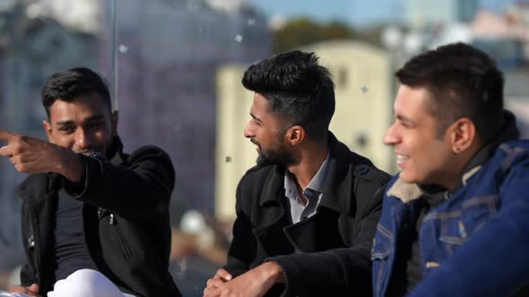 Positive Young Carefree Middle Eastern Men Talking Smiling Sitting at Glass Fence on Rooftop alt
