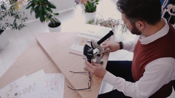 Male Teacher Using Microscope and Looking at Biology Textbook in Modern School alt