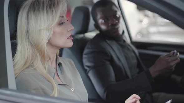 Side View of Beautiful Blond Caucasian Woman Sitting in Car Salon with Tablet and Explaining alt