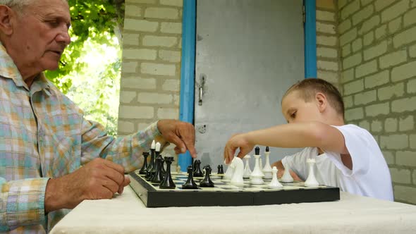 Grandfather and Grandson Play Chess in the Courtyard of the House
