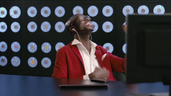 Black Man in Red Suit Sits at Desk Listens to Music Via Headphones and Dances