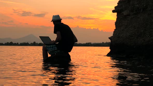 Programmer with Notebook Sitting on the Rock in the Sea on Sunset Background. Freelancer Working on