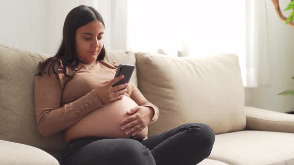 A young pregnant hispanic woman sitting on the couch with the smart phone alt