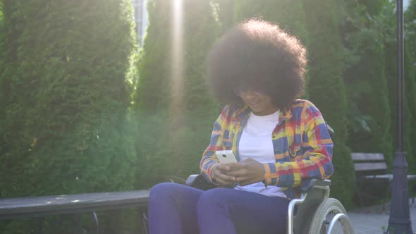 African American Woman with an Afro Hairstyle Disabled in a Wheelchair Uses a Smartphone Sunflare alt