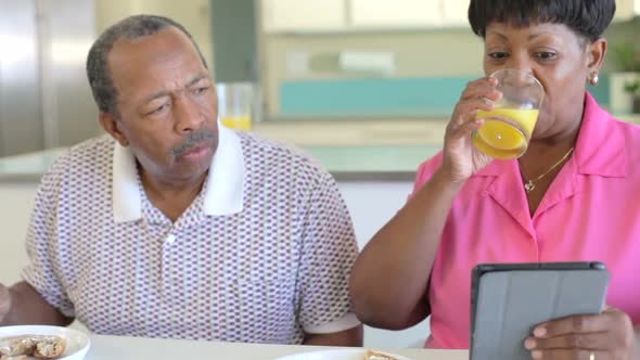 Senior Woman Using Digital Tablet As Husband Makes Breakfast 2 alt
