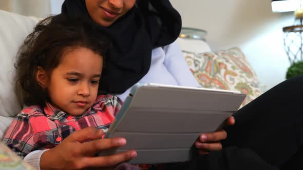 Young mother using a digital tablet with her daughter on the sofa at home 4k alt