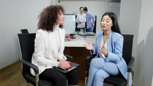 Positive Asian and Caucasian Women Talking and Shaking Hands in Office with Blurred Middle Eastern alt