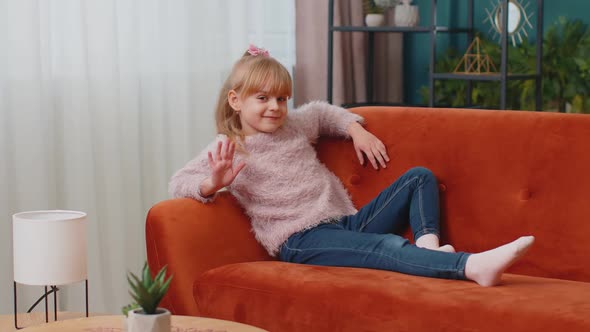 Child Girl Lying on Sofa at Home Looking at Camera Smiling Waving Hands Gesturing Hello or Goodbye alt