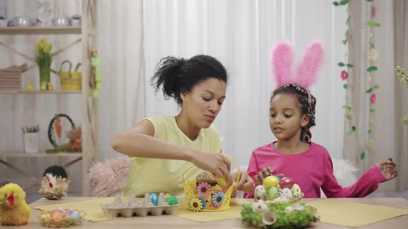 Mom and Daughter with Funny Bunny Ears Putting Easter Cake and Colored Eggs in Basket alt