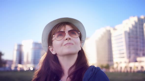 Happy Brunette Woman in a Hat Stands Against the Background of Houses and Looks Around