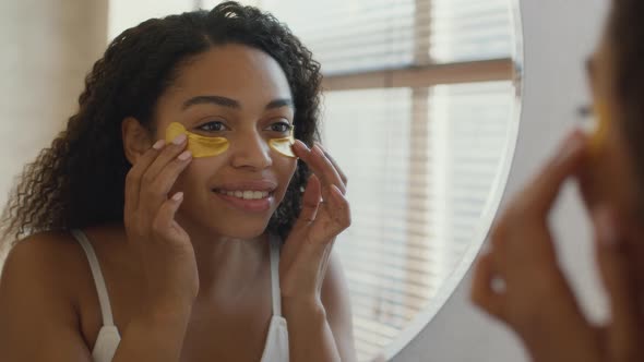 Young Pretty African American Woman Applying Anti Edema Collagen Patches on Face Looking at Mirror alt