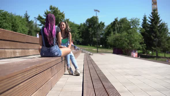Two Attractive Girlfriends Talking to Each Other Having a Good Mood Sitting on a Bench in the Park alt
