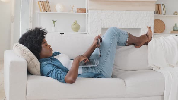 Smiling Serene African Young Woman Lying Down on Sofa Texting on Laptop alt