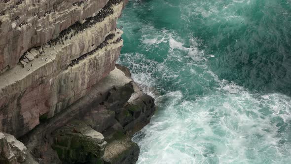Powerful waves crash over rocks and against a sea cliff in the middle of a beautiful, deep teal ocea alt