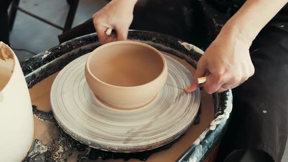Master is cutting clay cup from potter's wheel, close-up in workshop. She is using wire, and moving