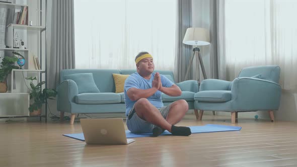Fat Asian Man On Mat Looking At Laptop And Doing Yoga In Sukhasana Pose At Home alt