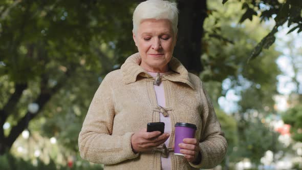Smiling Attractive Elderly Grayhaired Retired Woman Walking in Park with Plastic Cup of Coffee and alt