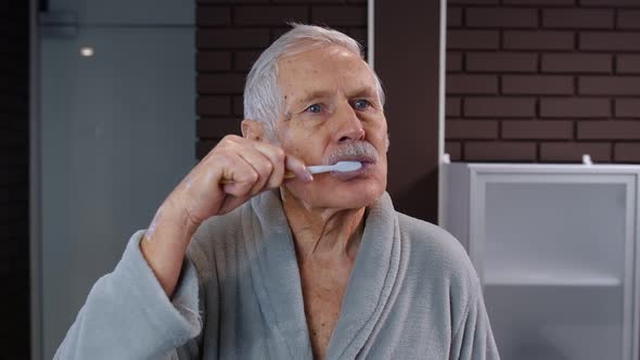Senior Man Grandfather in Bathrobe Brushing Teeth, Looking Into Mirror. Morning Hygiene at Bathroom alt