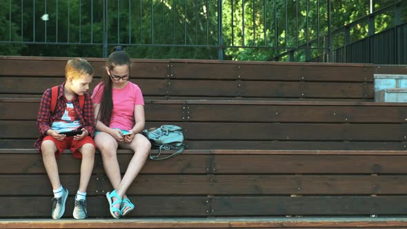 The Older Sister and Brother Are Sitting on the Street, Looking at Smartphones. alt