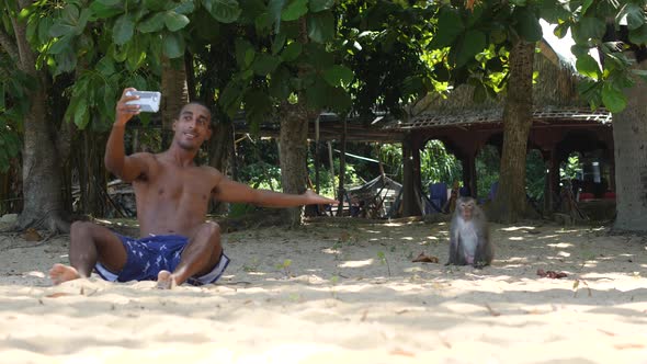 Tourist Man Sitting on the Sand Taking Selfie with the Monkey at the Beach. alt