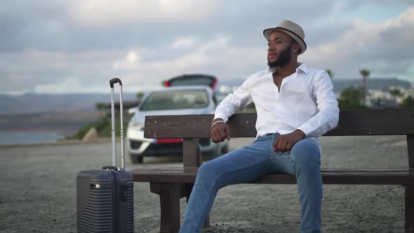 Wide Shot Portrait of Thoughtful African American Gay Man with Trolley Bag Sitting on Bench Looking alt