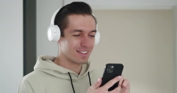 Closeup Portrait of Joyful Handsome Young Man Listening to Music in White Headphones and Surfing alt