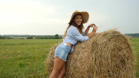 Pretty Young Woman Stands Near Haystack and Plays With Straw. Farmer's Wife Walks on the Field