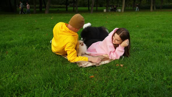 Young Guy and Girl Play with a Little Cute Dog Lying on the Grass in the Park alt