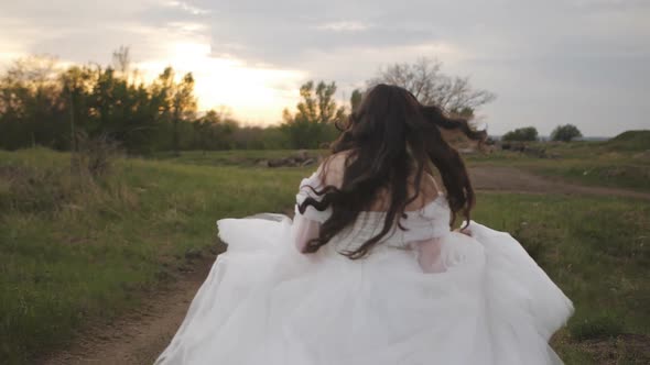 A Girl in a Wedding Dress and Her Hair Flying in the Wind Runs Along a Dirt Road a View From the