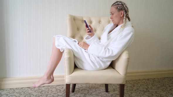 Young woman in white bathrobe uses mobile phone while sitting on armchair. alt