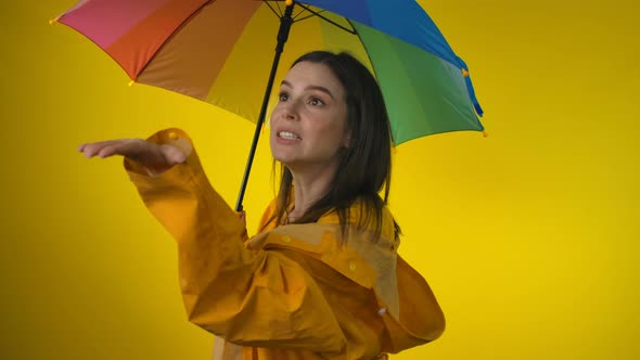 A Woman in a Yellow Raincoat with Rainbow Umbrella is Checking for the Rain alt