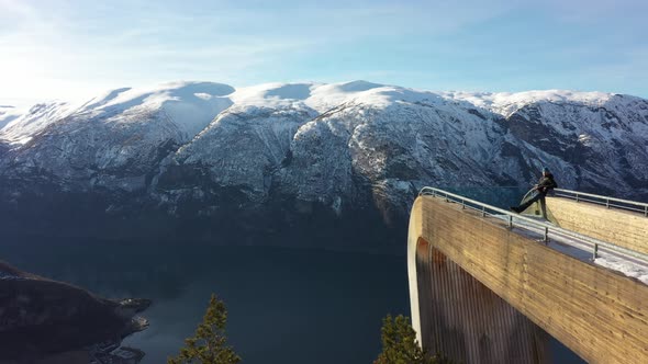 Tourist relaxing on tip of Stegastein viewpoint platform while drone slowlying along side and passin alt