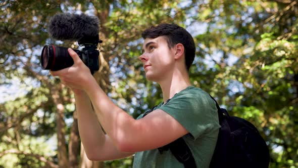 Close up of young male vlogger holding a professional camera shooting videos in a bright sunny fores alt