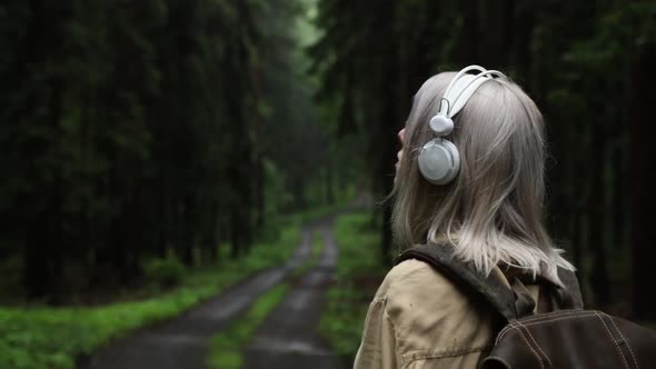 Blond hair woman in headphones with backpack in rainy forest