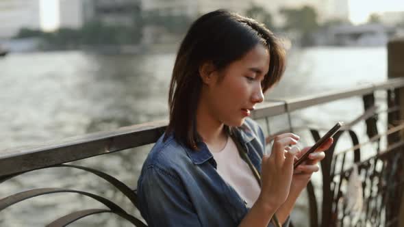 Young Asian female using smartphone while sitting beside Chao Phraya river in Thailand.