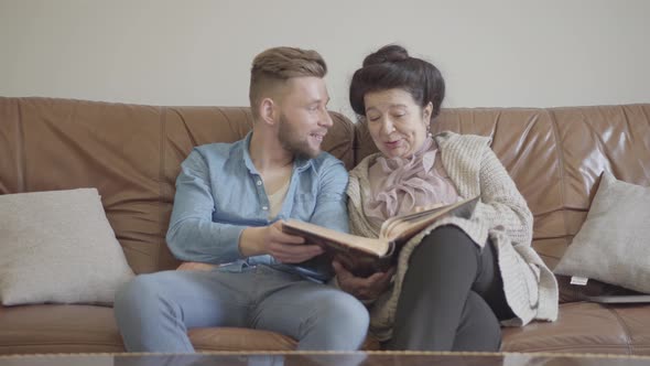 Cute Pretty Granny and Adult Grandson Sitting at Home on the Brown Leather Sofa Watching Old Photos alt
